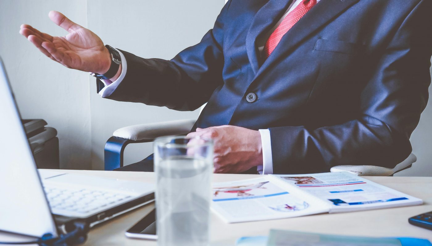 homme en costume main levé assis sur un bureau devant son ordinateur